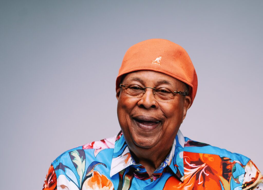 Close-up portrait of a smiling man wearing an orange Kangol cap and a colorful patterned shirt.